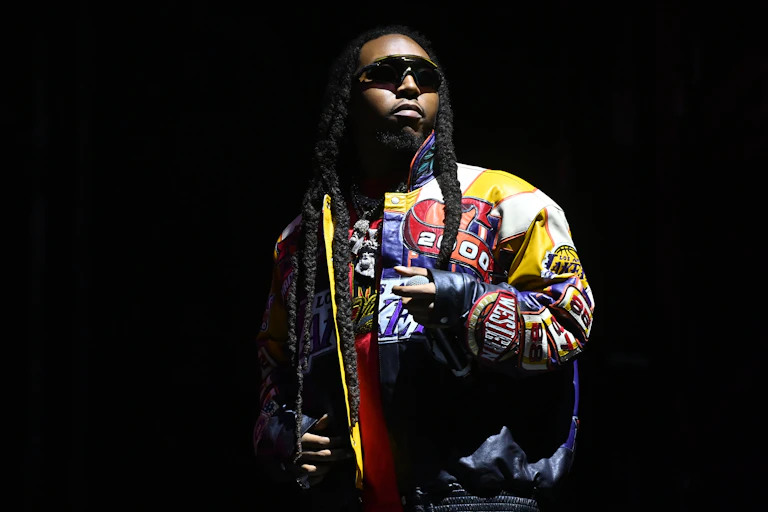 A young black man wearing dark sunglasses stands in the light on a dark stage.
