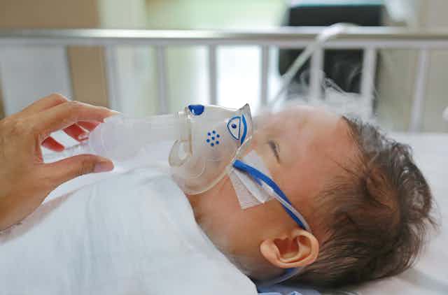 A toddler boy lies on his back with a nebulizer over his nose and mouth and mist rising from the inhaler.