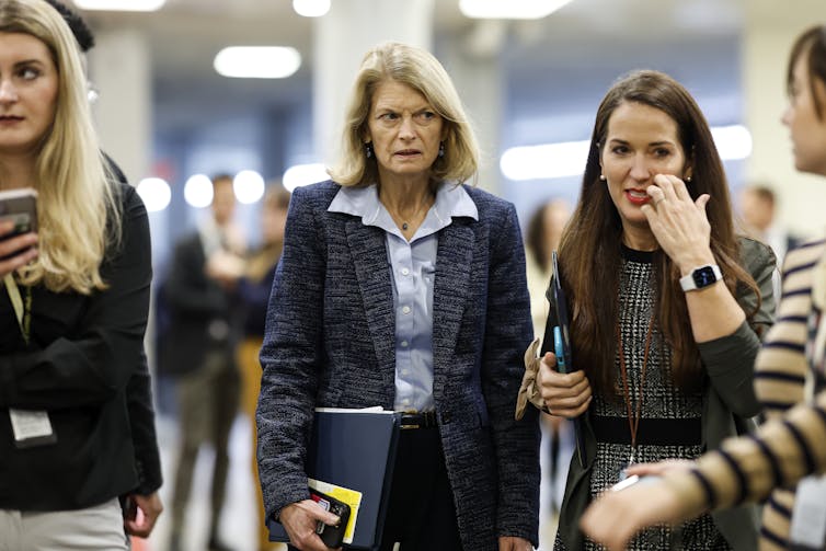 A middle aged white woman with blonde hair and a blue pantsuit walks through a room, with other younger women at her side.