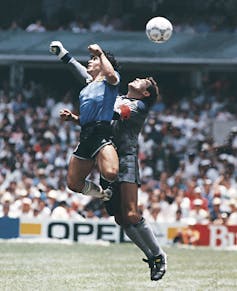 A soccer player in a blue jersey out-jumps a goalkeeper. A football flies over their heads.