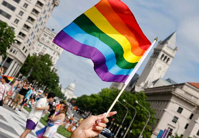 A hand is seen holding up a rainbow flag, with a crowd of people behind in a city street. The US Capitol is seen in the background.