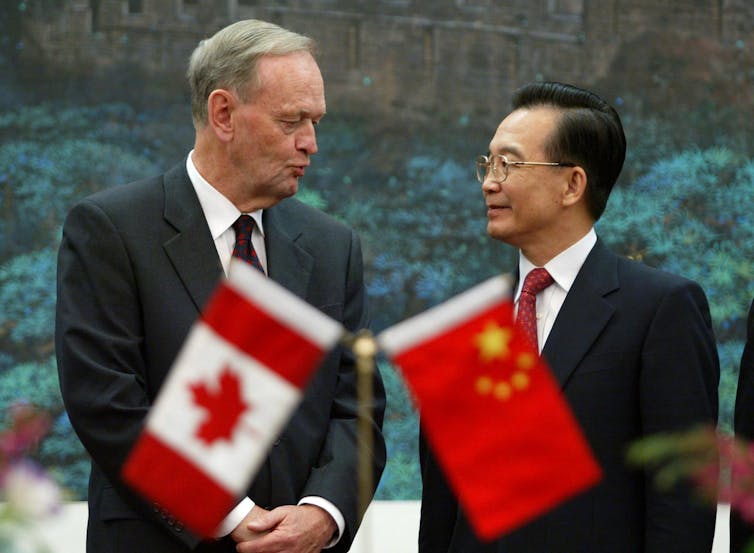 Two men in suits chat. A Canadian flag is in front of one and the Chinese flag is in front of the other.