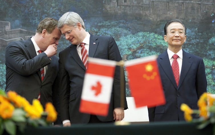 A dark-haired man whispers into the ear of a grey-haired man with glasses as a Chinese man stands next to them. The Canadian and Chinese flags are seen in front of them.