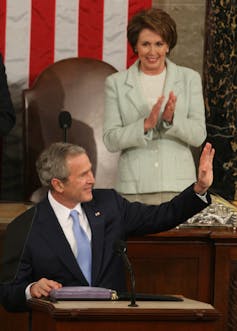 A woman in a light jacket stands behind a man in a suit as he waves.