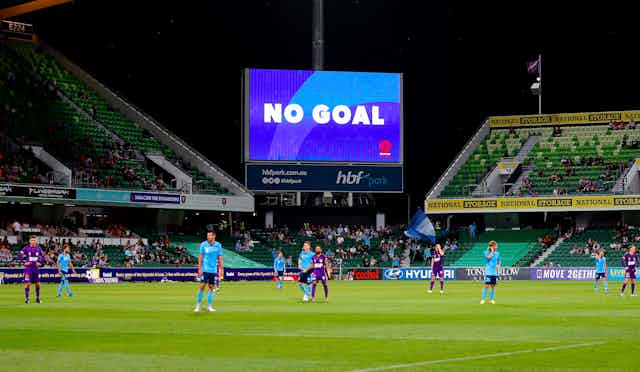 Players stand dejectedly on a football field while the scoreboard screen reads NO GOAL above them.