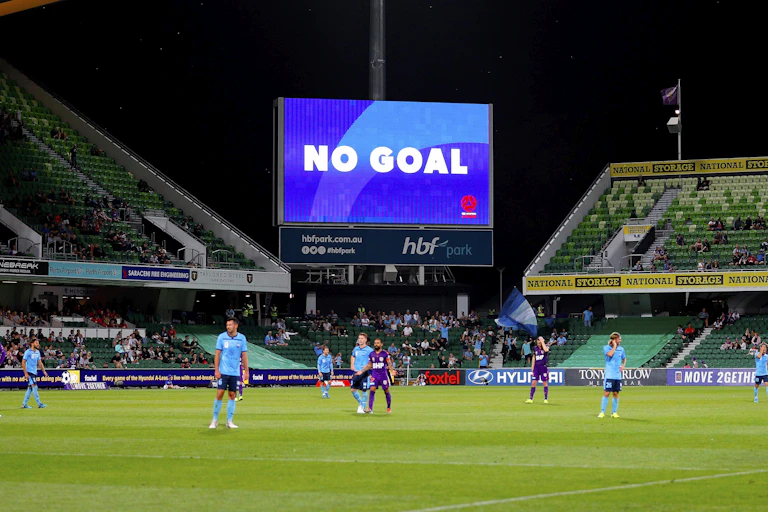 Players stand dejectedly on a football field while the scoreboard screen reads NO GOAL above them.