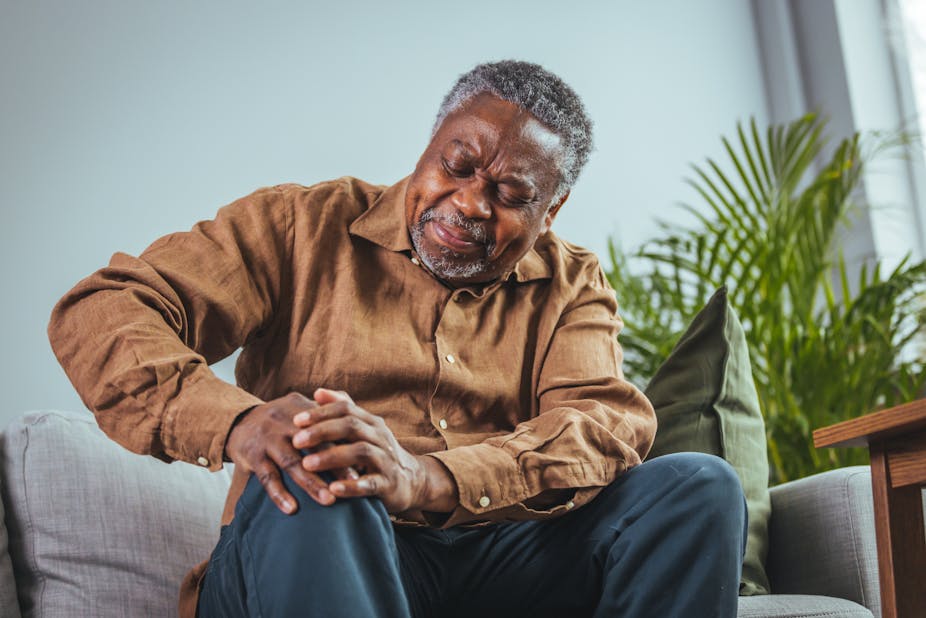 An older man of African ethnicity sits on a couch holding his knee, grimacing in pain. He is wearing dark blue trousers and a camel-coloured shirt