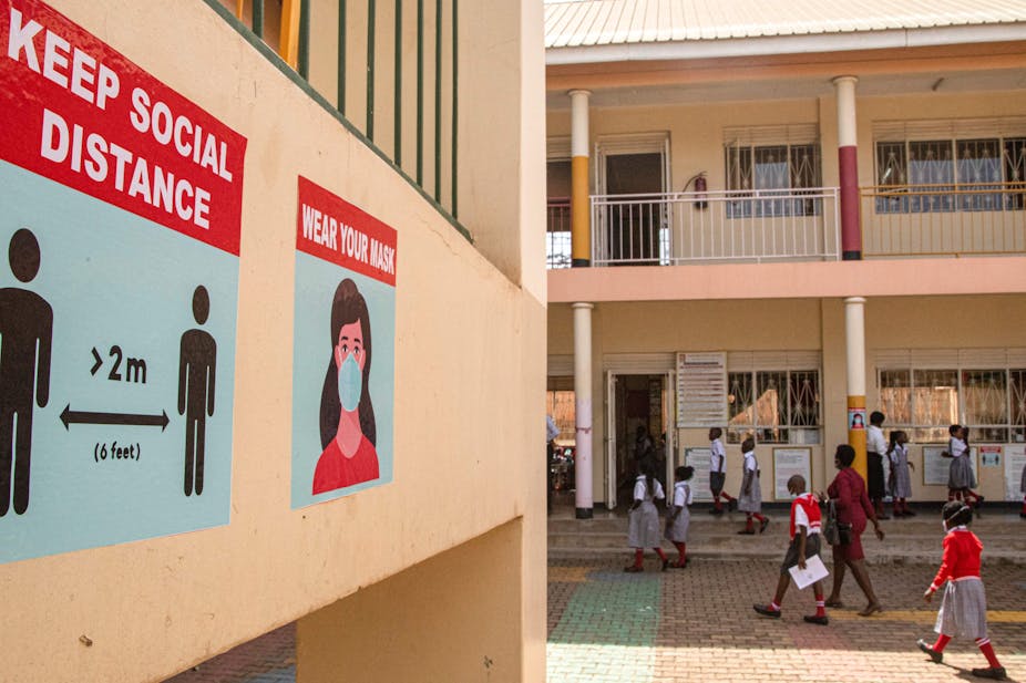 Children in school grey, red and white school uniforms walk past a wall painted orange-pink on which posters calling for social distance and mask-wearing are featured