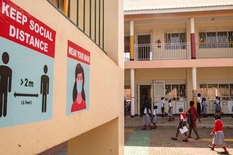 Children in school grey, red and white school uniforms walk past a wall painted orange-pink on which posters calling for social distance and mask-wearing are featured