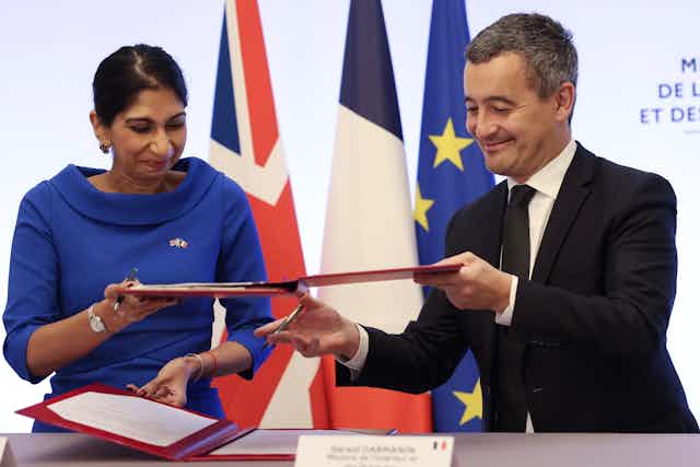 Suella Braverman and French Interior Minister Gerald Darmanin sign papers in red folders, standing in front of UK, France and EU flags