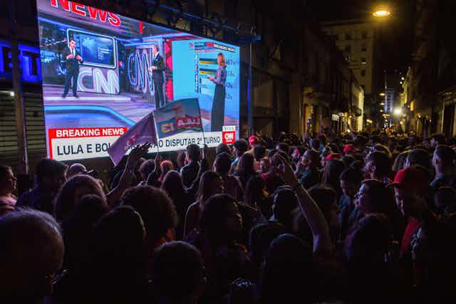 People standing in front of a big screen showing the Brazilian election.
