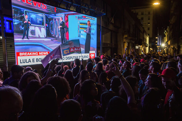 People standing in front of a big screen showing the Brazilian election.