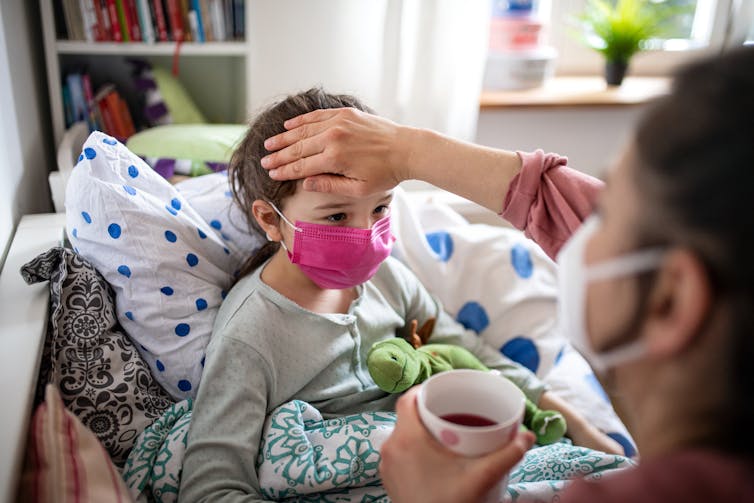 A woman feels her daughter's forehead. Both wear masks and the girl is in bed.