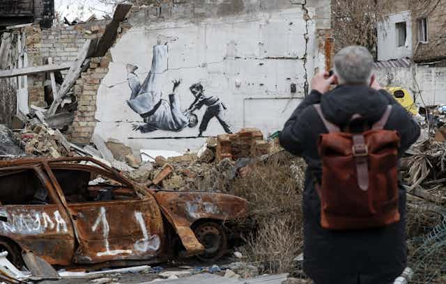 A man with a backpack faces away from us, taking a photograph of the Banksy work stencilled on the wall behind him. On the left is a burned out car. Behind it, the wall of a bombed building with Banksy's image of a young boy in Judo gear throwing down Putin