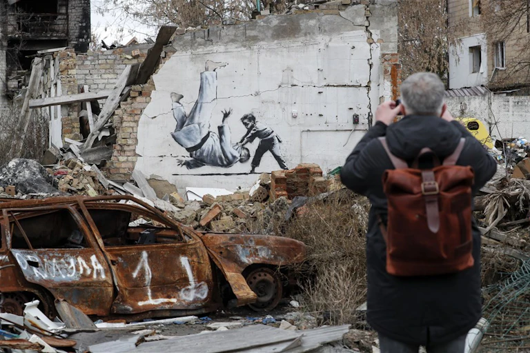 A man with a backpack faces away from us, taking a photograph of the Banksy work stencilled on the wall behind him. On the left is a burned out car. Behind it, the wall of a bombed building with Banksy's image of a young boy in Judo gear throwing down Putin