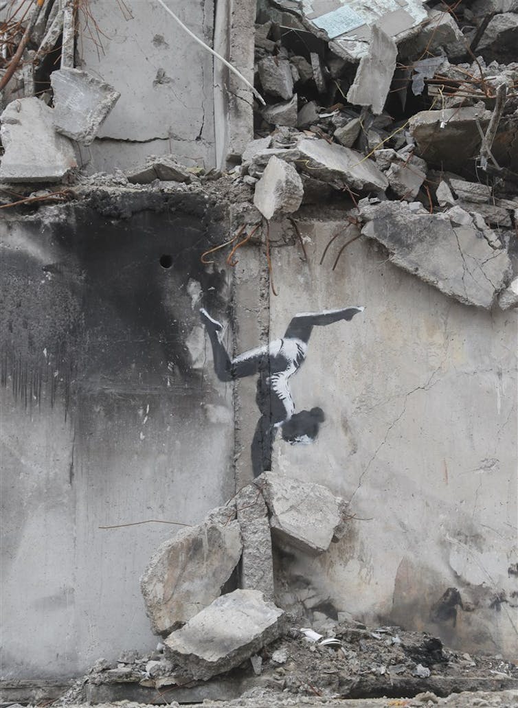 A ruined building with a Banksy mural on its walls, a ballerina doing a headstand on a pile of rubble beneath her