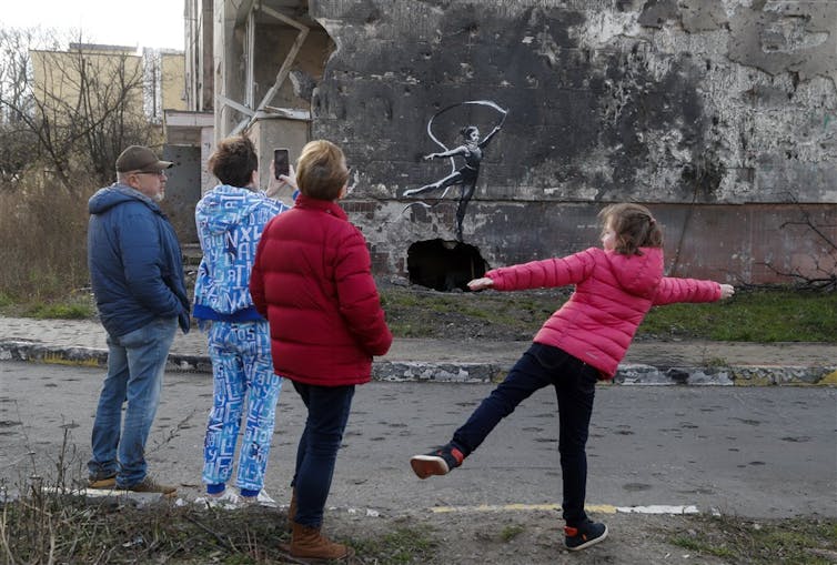A family looks at mural of a gymnast twirling a ribbon on the wall of a bombed building, girl mirrors the gymnast's pose