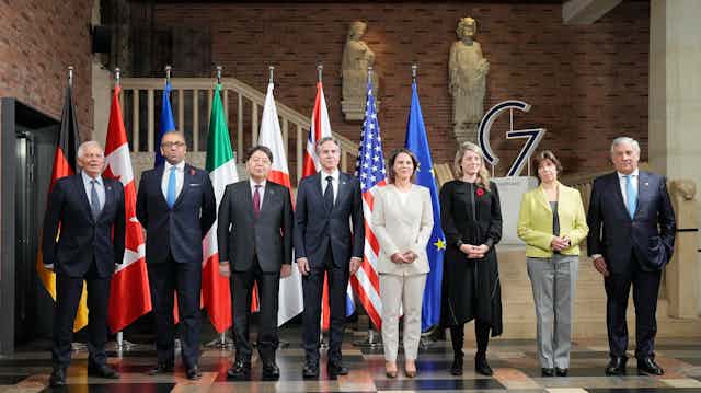 Five men and three women pose in front of a row of flags from the G7 nations