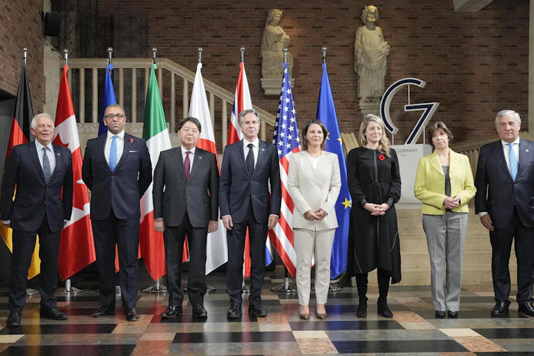 Five men and three women pose in front of a row of flags from the G7 nations