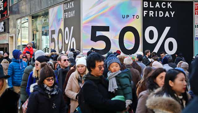 crowds of people, some wearing stocking hats, walk in the street past big black friday signs advertising 50% offf