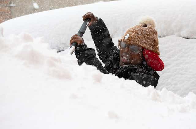 A woman shovels snow off her car, not visible under the piles.