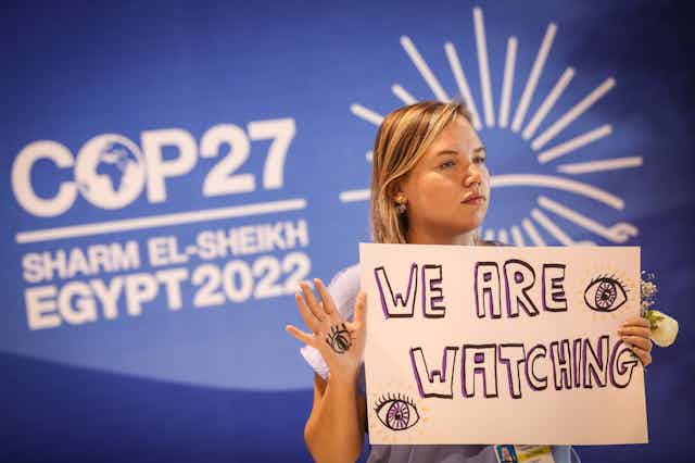 A young person holds a sign saying 'we are watching' in front of a display with COP27 on it.