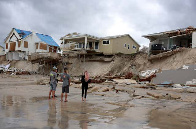 People stand on the beach looking up at home where erosion has cut beneath them. One has partially collapsed, another has deck and pool suspended precariously. They once had ground beneath them.