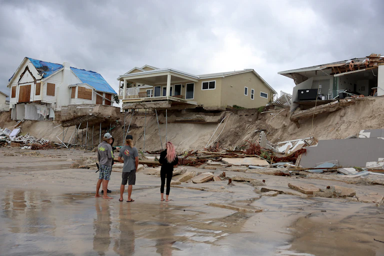 People stand on the beach looking up at home where erosion has cut beneath them. One has partially collapsed, another has deck and pool suspended precariously. They once had ground beneath them.