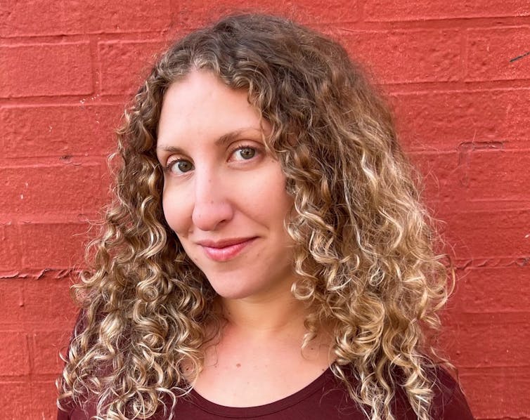 A smiling woman with curly hair in front of a painted red brick wall.