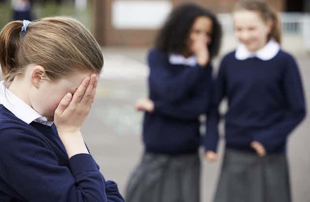 Two school students laugh at another student, with her head in her hands.