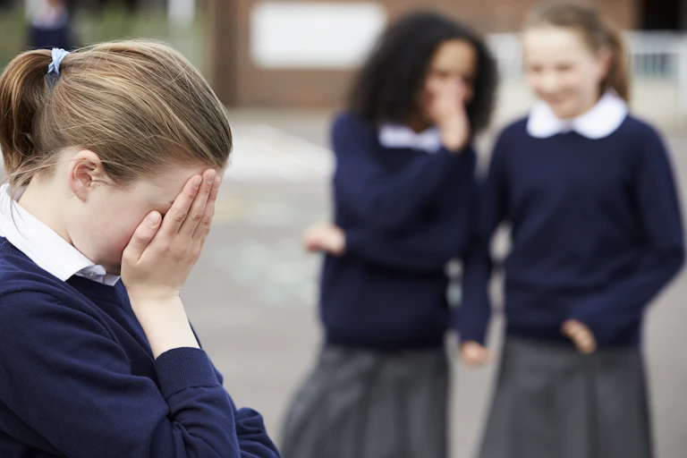 Two school students laugh at another student, with her head in her hands.