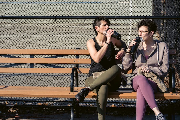 Two women sit on a bench, sipping water