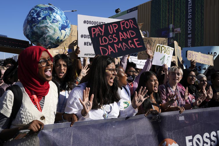 Young people from many countries shout and wave signs reading 'pay up for loss and damage' at a small outdoor protest.
