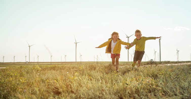 Two young children holding hands and running in a sunny field in front of wind turbines