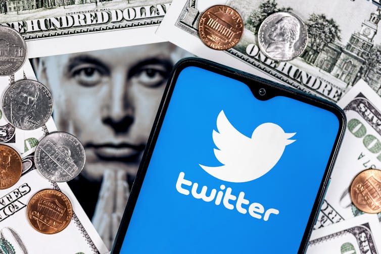 a phone screen showing the twitter logo of a silhouette of a white bird on a blue background sits atop a pile of money and a photograph of elon musk