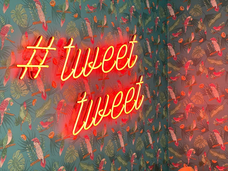 neon sign in red reading #TWEET TWEET, installed on top of green patterned wallpaper