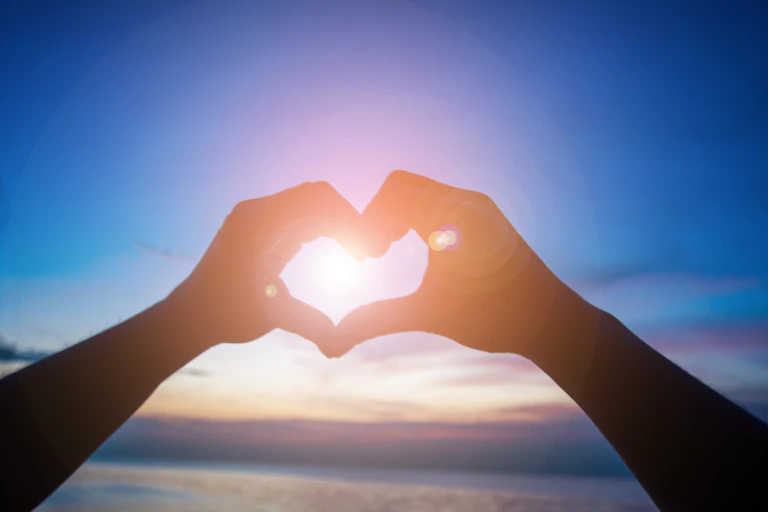 Dos manos dibujan un corazón contra el sol de la tarde.
