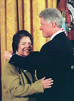 A tall white man with thick gray hair places medal around neck of shorter woman with cropped brown hair