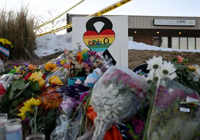 A sign showing a rainbow heart and 'club Q' on it sits atop flowers and in front of police 'do not cross' tape.