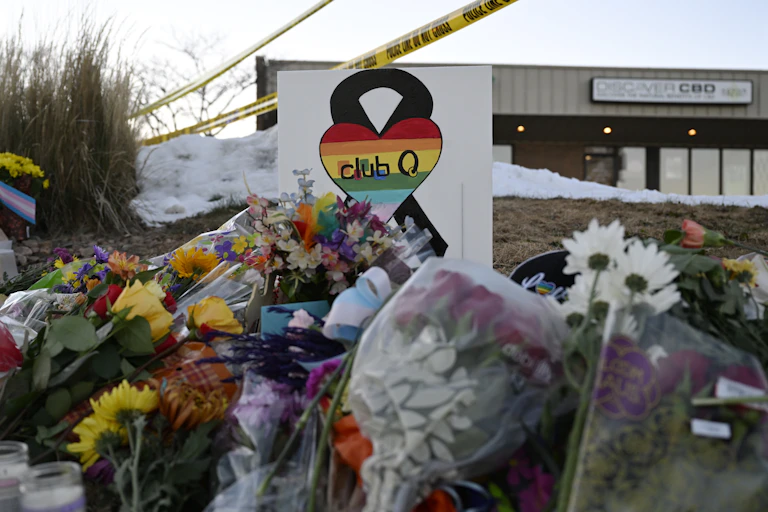 A sign showing a rainbow heart and 'club Q' on it sits atop flowers and in front of police 'do not cross' tape.