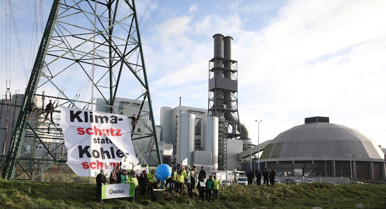A group of environmental activists standing in front of a coal power station, with policeman grouped to the side.