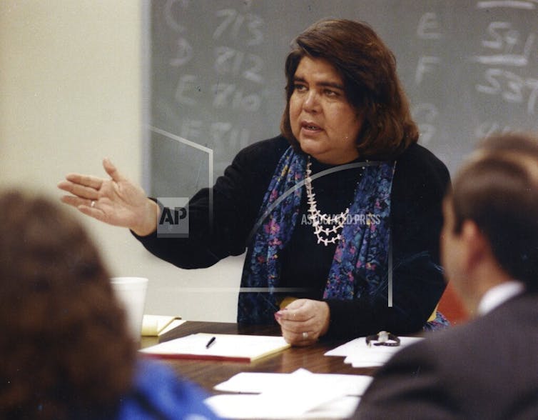 woman with thick brown hair sitting at a desk in a classroom and outstretching her arm