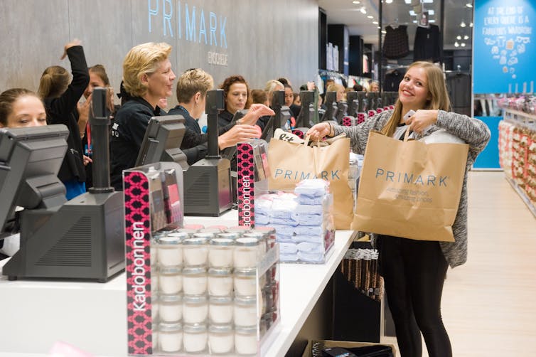 Smiling woman with blonde hair lifts two large Primark paper bags from a counter of a Primark shop.
