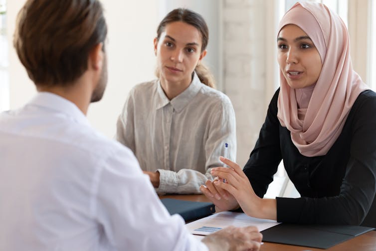 A man sits facing away from the camera, talking to two women at a desk. One of the women is wearing a hijab.