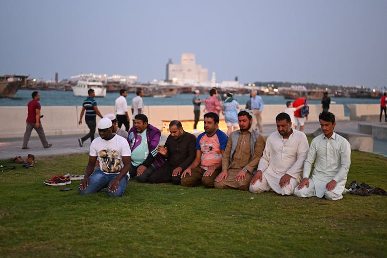 A row of middle aged, brown skinned men sit on their knees in a line on grass, with water in the background.