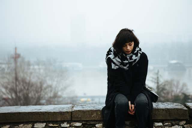 A sad young woman sits on a stone fence. A snowy winter scene is in the background.