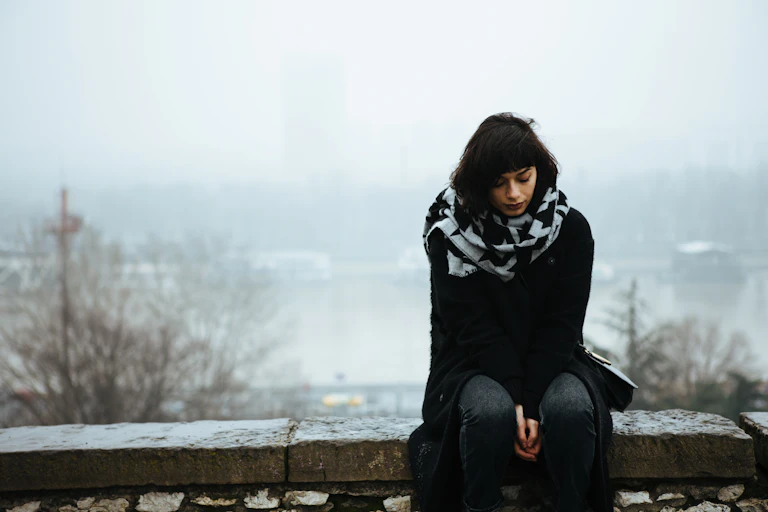 A sad young woman sits on a stone fence. A snowy winter scene is in the background.