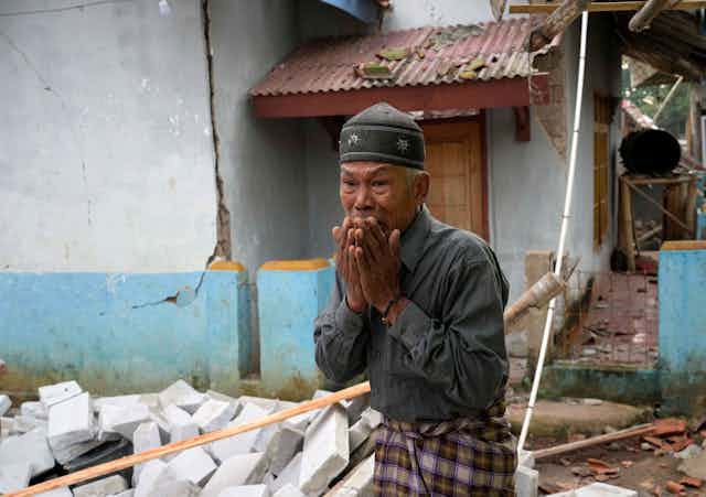 A distraught elderly man with ruined buildings in the background