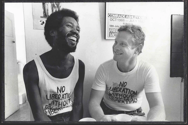 Two men sit at a table with coffee mugs in front of them, smiling. Behind them is a political poster.