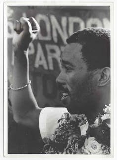 A man holds up his fist, a garland of flowers around his neck, a banner behind him.